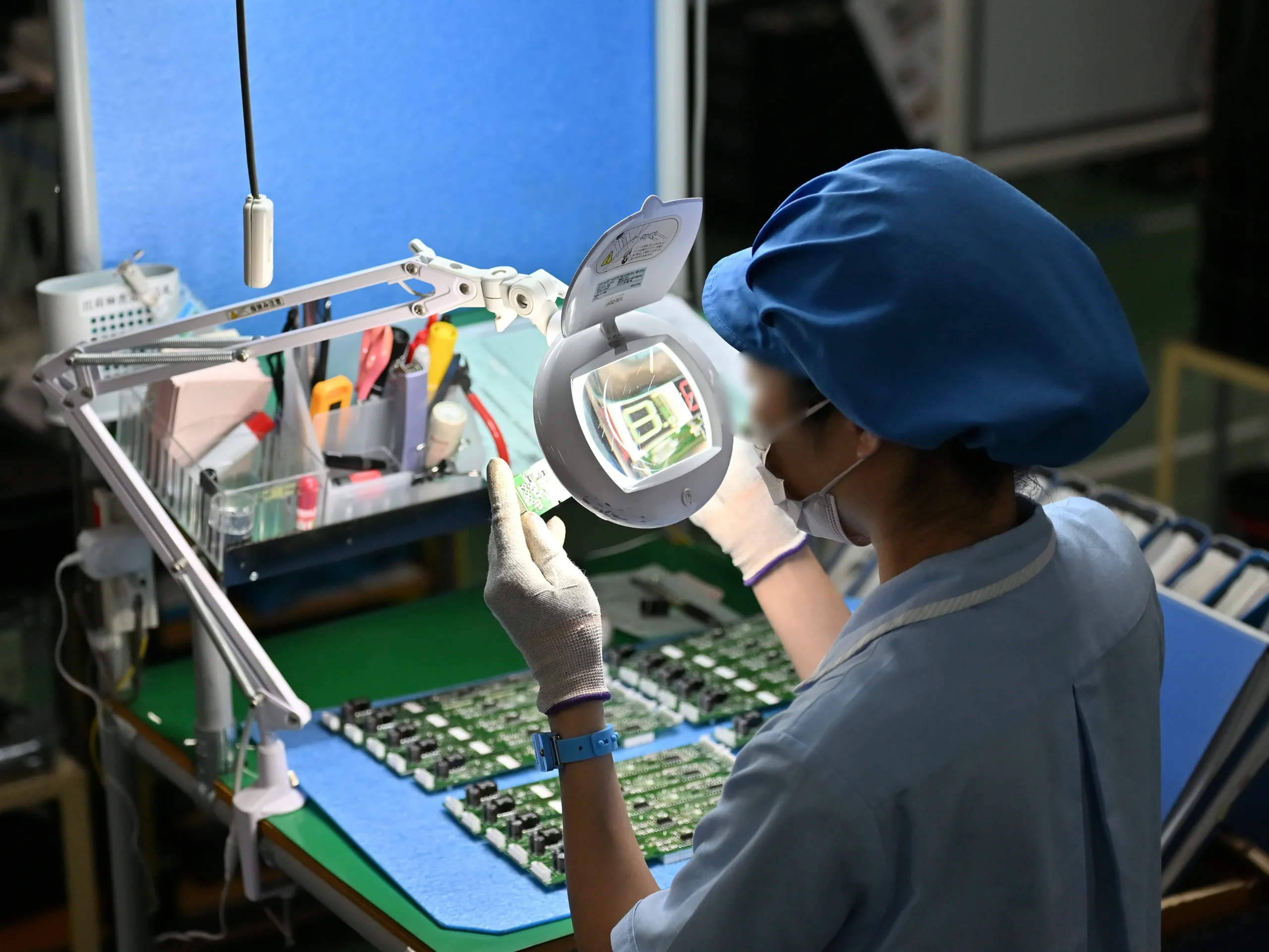 青い帽子をかぶった作業員が、拡大鏡付きライトの下で電子基板を精密に検査しています。従業員約150人から年間4,768件の改善提案が生まれる5S活動は、製造現場だけでなく、フリーアドレス化に不可欠なクリアデスクを推進する事務所でも実践されています。