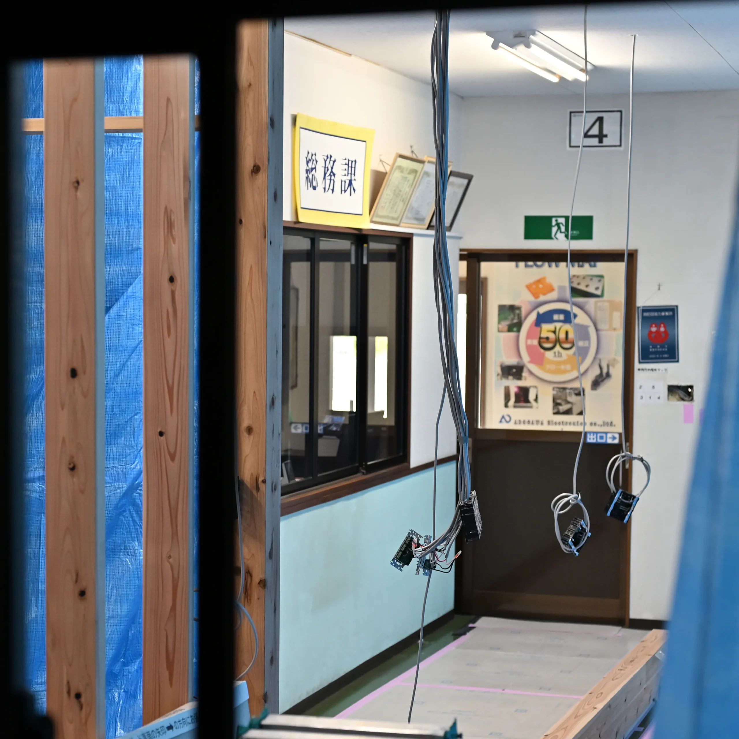 事務所改装工事中の廊下。左側に木材の骨組みと青い養生シート、天井からは電気配線が垂れ下がっている。改装工事は、フリーアドレス化によるコミュニケーション活性化やスペース効率化を目的としている
