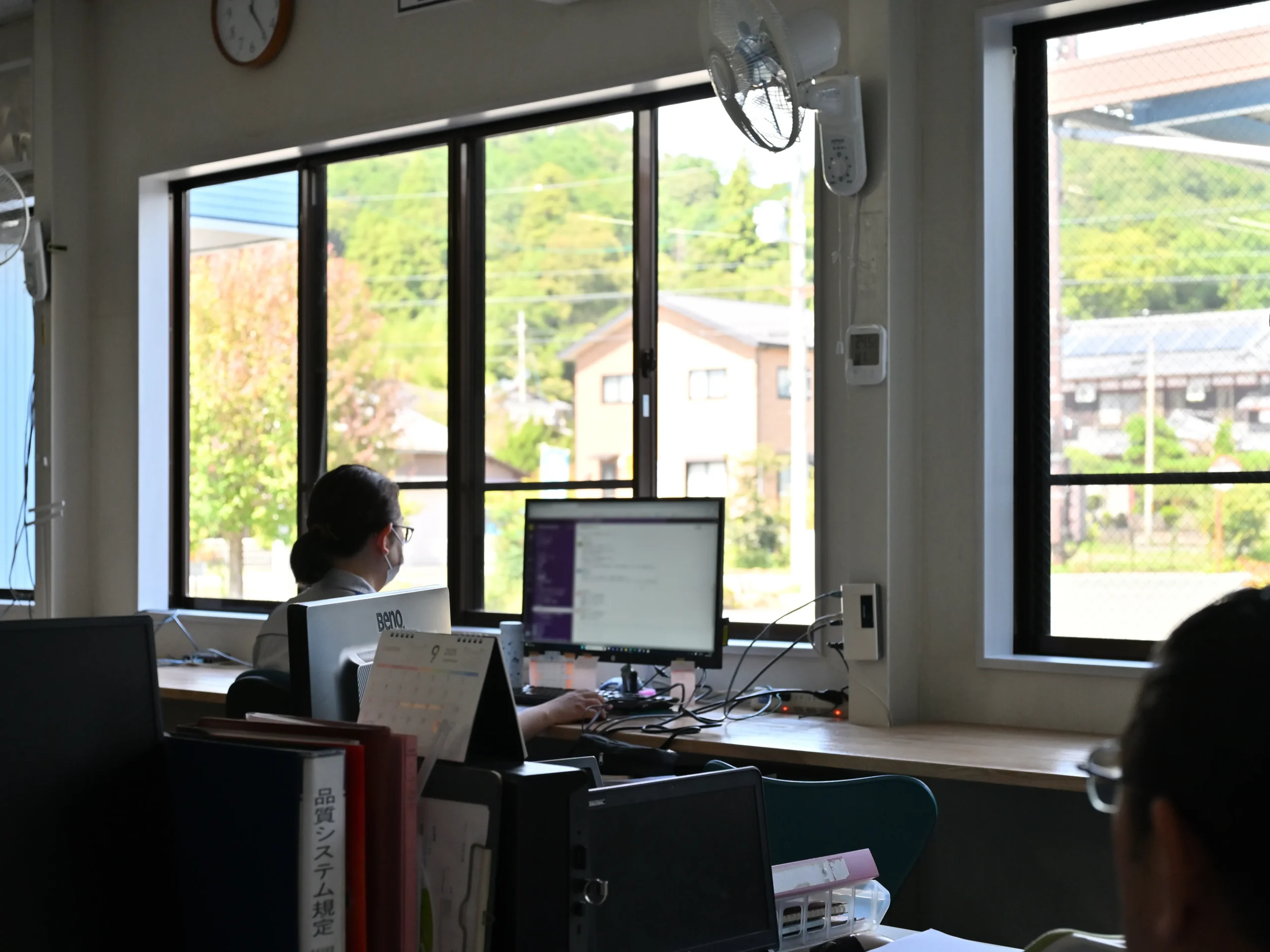 緑豊かな窓の外を望むオフィスで、従業員がデスクワークをしています。全社で年間4,768件の改善提案を生む5S活動は事務所でも実践され、クリアデスクの徹底が情報セキュリティの強化にも貢献しています。