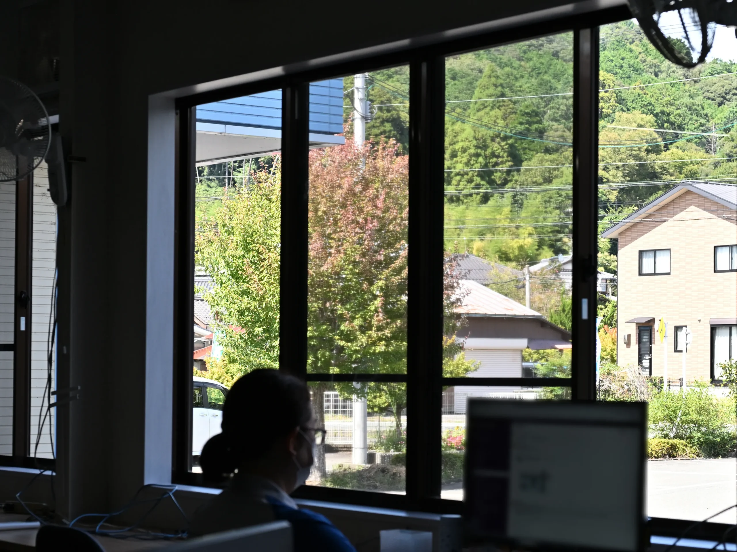 改装工事中のオフィスで、窓際のテーブルに座ってパソコン作業に集中する従業員。フリーアドレス化を進める中、打ち合わせ用テーブルの搬入や配線整備が行われている。大きな窓からは住宅街の緑と電線が見え、季節の移ろいを感じる秋の風景が広がっている。