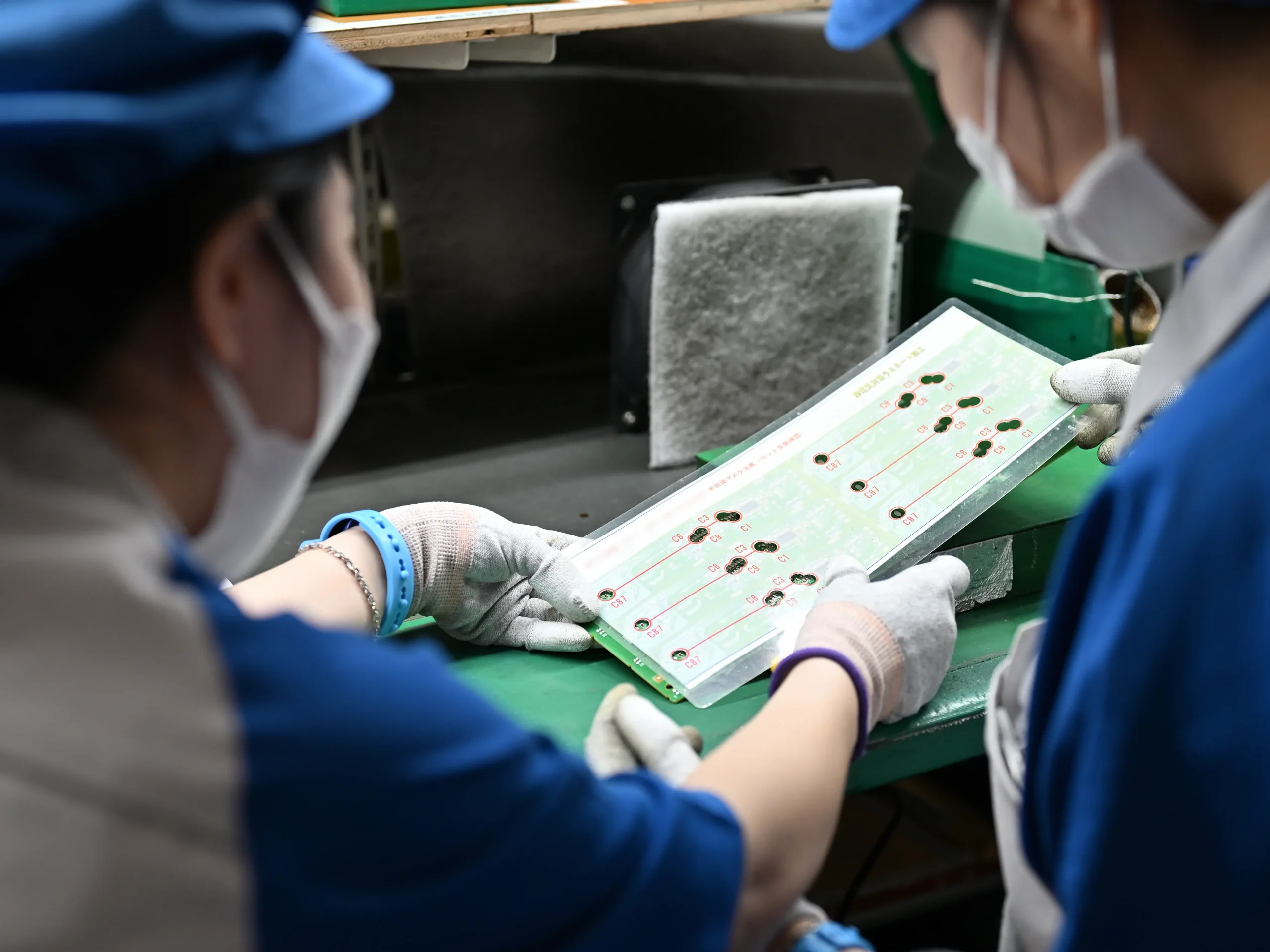 電子基板を検査する従業員の様子。青い作業着と白い手袋を着用し、品質管理を徹底する姿が映されている。2024年10月〜2025年8月の11か月間で、約150名の従業員による5S・改善活動の年間提案件数は4,768件に達し、1人あたり月平均2.89件の改善提案が実施された。