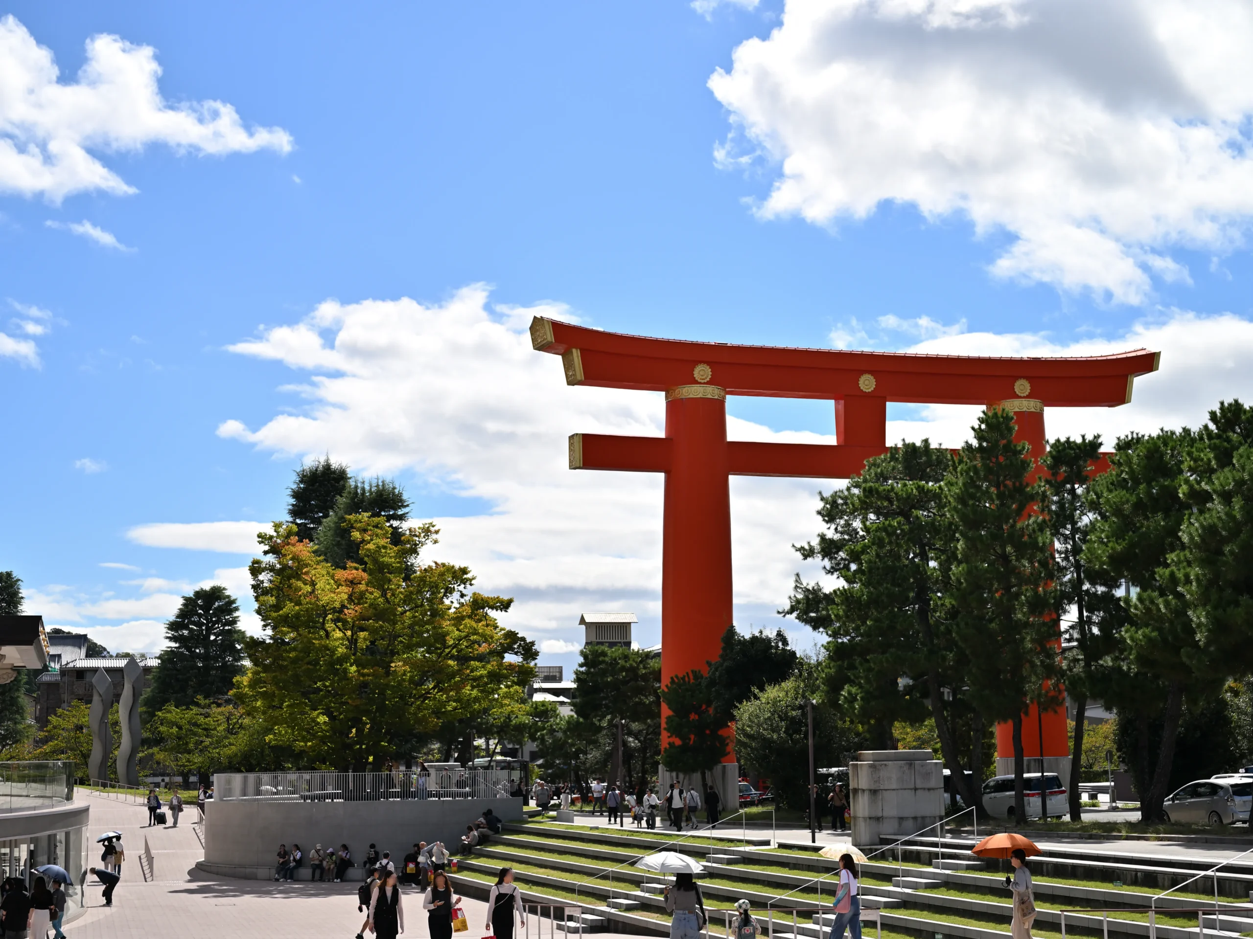 緑の木々と朱色の巨大鳥居(平安神宮)、京都市京セラ美術館の階段状の広場を行き交う人々。