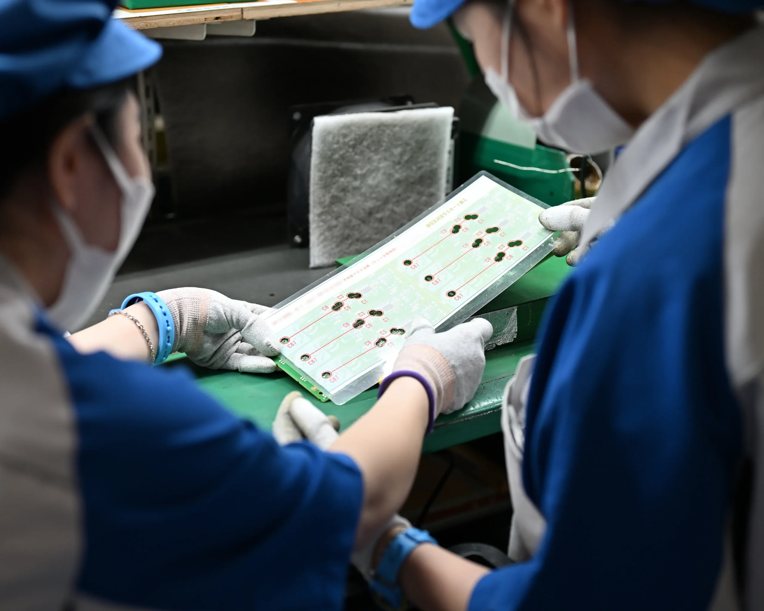 プリント基板のはんだ付け目視検査の方法やコツを、同僚に丁寧に説明する様子。業務中のコミュニケーションが、健全な社内風土づくりの方法とやる気を持続させる組織文化のポイント。