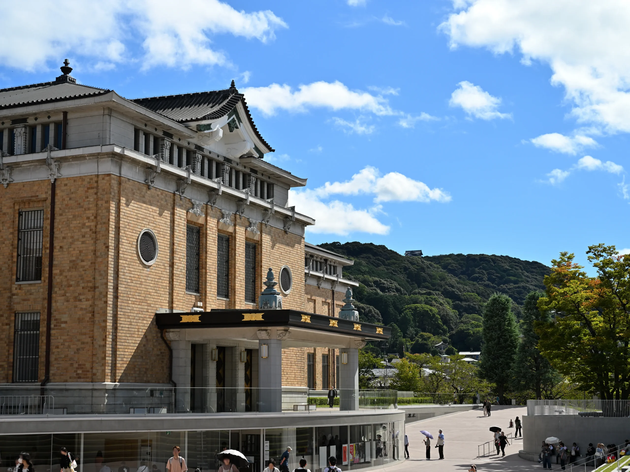 晴れた日の京都市京セラ美術館の外観と周辺の広場。煉瓦タイルの建物と青空、緑の山。